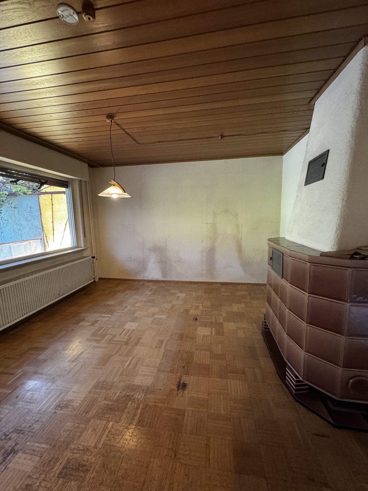 Vorher: leerer Wohnraum mit Holzdecke, Kachelofen und fleckigen Wänden